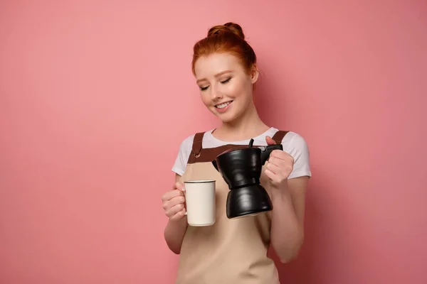 Den rödhåriga flickan i ett förkläde står i en halvsväng på en rosa bakgrund, lutar kaffekannan till koppen — Stockfoto