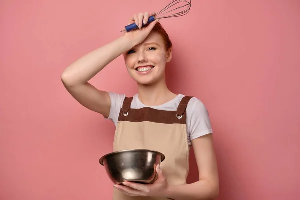 앞치마 안에 있는 붉은 머리 소녀는 분홍색 배경에 접시를 들고 손으로 이마를 맞대고 웃으며 서 있다 — 스톡 사진