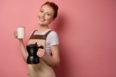 Önlüklü kızıl saçlı bir kız pembe bir arka planda, elinde kahve fincanı ve beyaz bir fincanla ayakta duruyor.