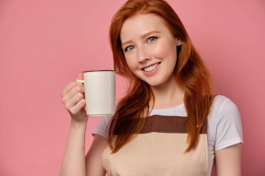 Önlüklü güzel kızıl saçlı kız pembe bir arka planda duruyor ve yüzüne beyaz bir kupa dayayıp gülümsüyor..