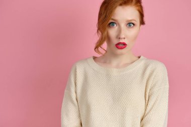 Kırmızı dudaklı, beyaz kazaklı kızıl saçlı bir kız pembe arka planda gözleri ve ağzı açık duruyor.