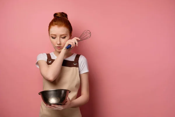 En rödhårig flicka i ett förkläde står på en rosa bakgrund, tittar in i en skål i handen och torkar av mjölet från ansiktet — Stockfoto