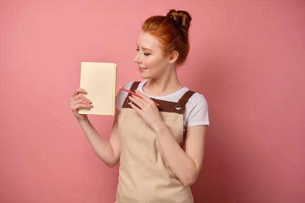 En rödhårig flicka i ett förkläde med insamlat hår står på en rosa bakgrund och pekar med en penna på sidan i anteckningsboken — Stockfoto