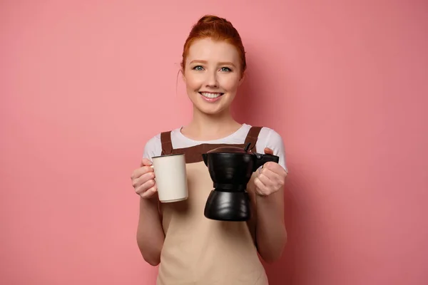 Rödhårig flicka i ett förkläde står på en rosa bakgrund med en kaffekanna och en kopp söt leende. — Stockfoto