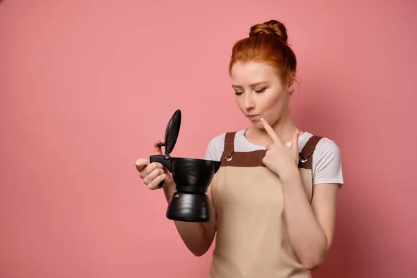 En rödhårig flicka i ett förkläde står i en halvsväng på en rosa bakgrund och ser tankfullt in i den öppna kaffekannan i handen — Stockfoto