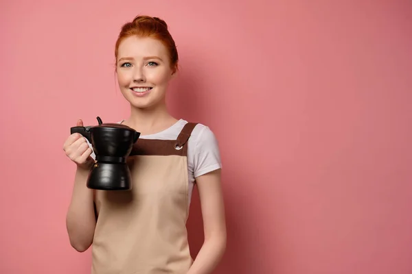 Vöröshajú lány összegyűjtött haj, kötényben, áll a rózsaszín háttér egy kávéskanna — Stock Fotó