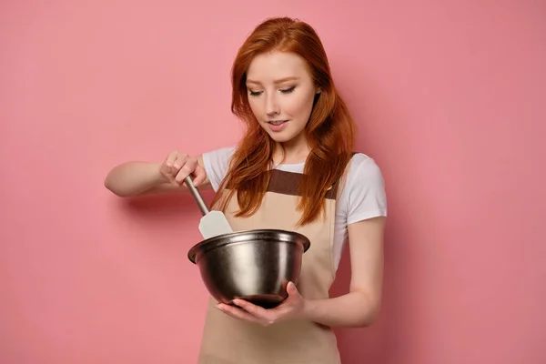 Den rödhåriga flickan i ett förkläde står på en rosa bakgrund och blandas med en spatel i en skål — Stockfoto