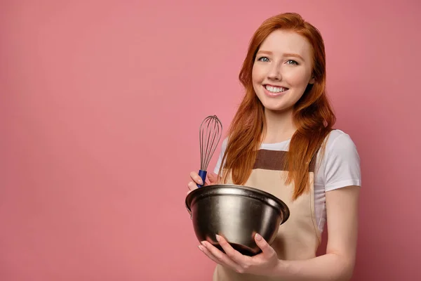 Den rödhåriga flickan i ett förkläde står på en rosa bakgrund och ler brett mot kameran med en kopp och en visp i händerna — Stockfoto