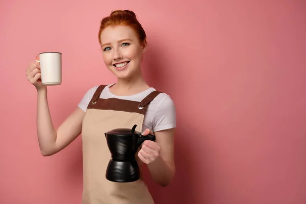 Rödhårig flicka i ett förkläde står i halvsväng på en rosa bakgrund med en kaffekanna, håller upp en kopp — Stockfoto