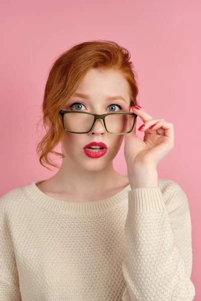 En rödhårig tjej i vit tröja med rött läppstift står på en rosa bakgrund, justerar glasögon och tittar på kameran — Stockfoto