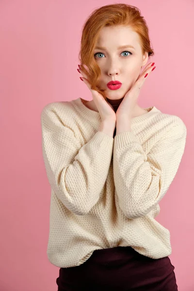 하얀 스웨터를 입고 붉은 립스틱을 바른 빨간 머리 소녀가 손을 얼굴에 대고 핑크 색 배경에 서 있다. — 스톡 사진