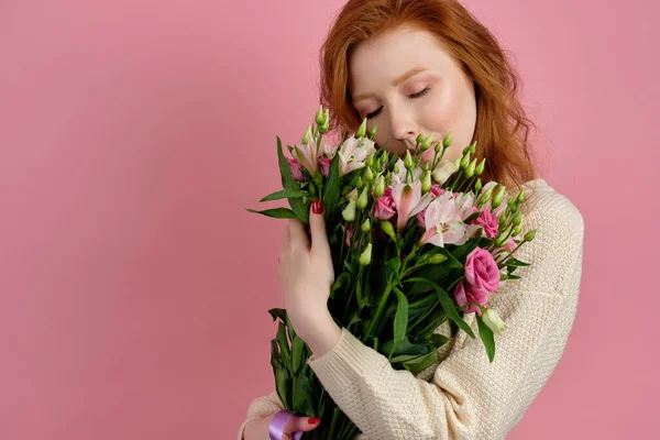 Egy vörös hajú, göndör hajú, fehér pulóveres lány áll rózsaszín háttéren, egy csokor virággal a kezében, és becsukja a szemét. — Stock Fotó
