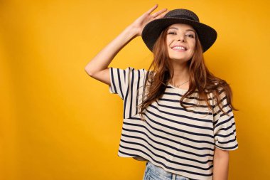 Hasır şapkalı güzel bir esmer sarı arka planda duruyor ve geniş bir şekilde gülümsüyor, çenesini kaldırıyor ve başının arkasından elini tutuyor.
