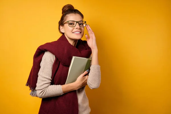 En vacker brunett med en hög bulle står i en tröja och halsduk, håller en bok och tittar på kameran med ett leende — Stockfoto