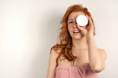 Kırmızı saçlı kıvırcık saçlı, pembe havluyla beyaz arka planda duran, gözünü krema kavanozuyla kaplayan bir kız.