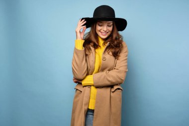 Sarı kazaklı ve ceketli bir esmer mavi bir arka planda duruyor, gülümsüyor, aşağı bakıyor, şapkasını eliyle tutuyor.