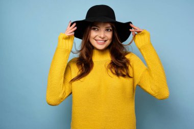 Sarı kazaklı bir esmer mavi arka planda duruyor, güzel gülümsüyor ve şapkayı elleriyle tutuyor.