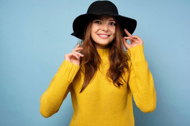 Sarı kazaklı ve şapkalı güzel bir esmer mavi arka planda duruyor, gülümsüyor, saçlarını düzleştiriyor.