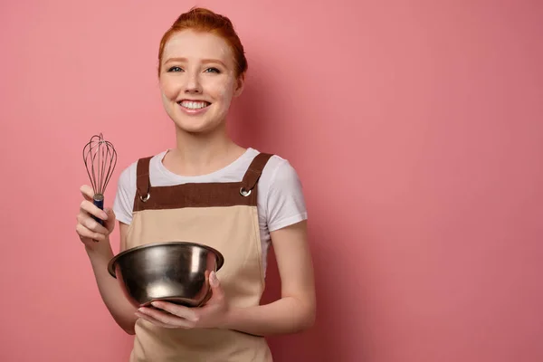 Den rödhåriga flickan i ett förkläde, målad i mjöl, står på en rosa bakgrund med en skål och en visp och leenden — Stockfoto