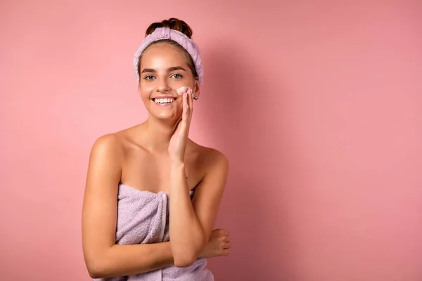 Seorang gadis dengan kulit bersih berdiri di atas latar belakang merah muda, tersenyum di depan kamera, menerapkan krim di wajahnya . — Stok Foto