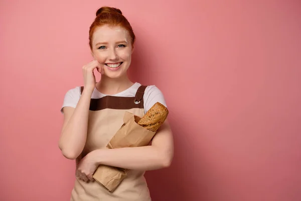 Vacker rödhårig flicka med samlat hår, i ett förkläde, med mjöl i ansiktet, ler, håller en påse bröd, med handen i ansiktet — Stockfoto