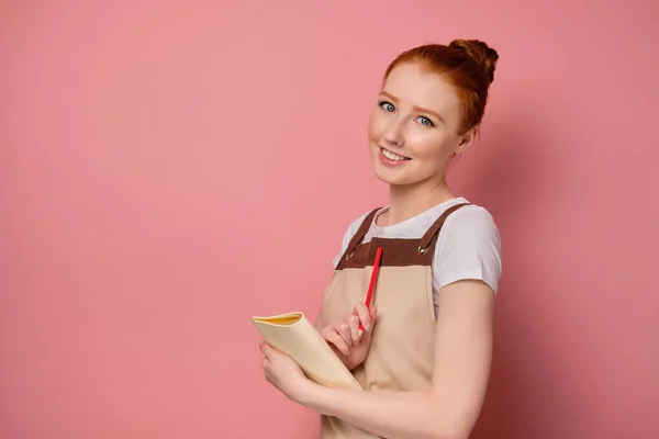 En rödhårig flicka i ett förkläde med insamlat hår står på en rosa bakgrund i halvtur med en anteckningsbok och ler gulligt. — Stockfoto