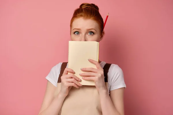 Egy vörös hajú lány egy kötényben, összegyűjtött hajjal, rózsaszín háttéren áll, a száját befedi egy jegyzetfüzettel és tágra nyílt szemekkel. — Stock Fotó