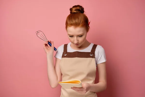 Den rödhåriga flickan i ett förkläde står på en rosa bakgrund, tittar in i en anteckningsbok i handen med en visp i handen — Stockfoto