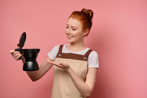 Egy vörös hajú, kötényes lány félfordulatban áll rózsaszín háttéren nyitott kávéskannával a kezében, és rámutat, mosolyogva. — Stock Fotó