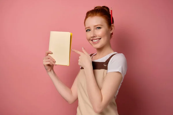 Egy vörös hajú lány egy kötényben, összegyűjtött hajjal, félkabátban áll, rózsaszín háttérrel, és ujjal mutogat a noteszre. — Stock Fotó