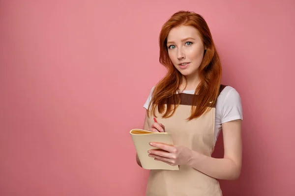 A red-haired girl in an apron stands on a pink background in half a turn, holds a notebook in her hands and looks at the camera — Stock Photo, Image