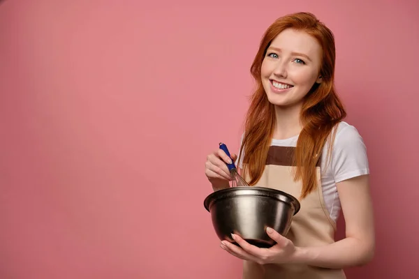 앞치마를 입은 빨간 머리의 여자 아이가 핑크 색 배경에 서서 채찍을 휘두르며 그릇에 수염을 물고는 환하게 틀에 미소를 짓습니다 — 스톡 사진