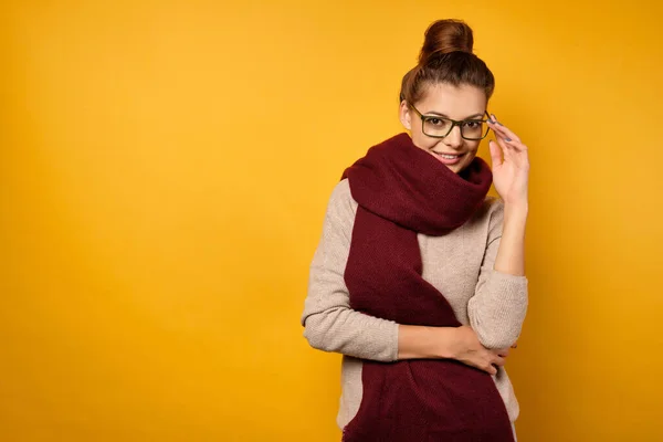 En vacker brunett med en bulle på huvudet och glasögon står i en tröja och, gömmer sig bakom en halsduk, tittar på kameran — Stockfoto