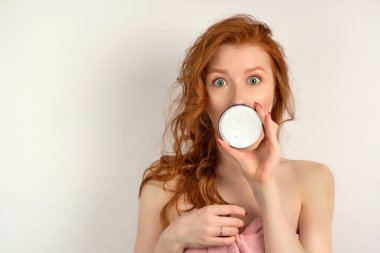 A red-haired girl with clean skin in a towel openseyes in surprise, looks into the camera, covering mouth with a jar of cream.