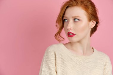 A beautiful red-haired girl with red lips in a white sweater stands on a pink background and, turning her head, looks to the side