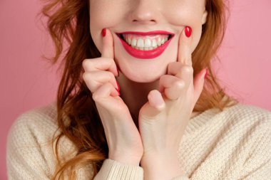 A close shot of the face of a red-haired girl with red manicure and lipstick, stretching a smile with fingers
