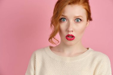Beautiful red-haired girl with red lips in a white sweater stands on a pink background in surprise opening her eyes and mouth