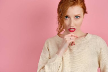 A beautiful redhead girl with red lips in a white sweater looks at the camera, holding chin with fingers