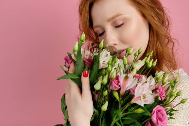 Beautiful red-haired girl gently hugs a bouquet of flowers, closing her eyes, on a pink background