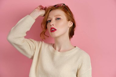 A beautiful red-haired girl with red lipstick putting away her glasses on forehead and looking at the camera with head back