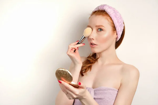 Red-haired girl with clean skin in a towel and a bandage on head with a mirror and closes eye with a brush, looking at the camera — Stock fotografie