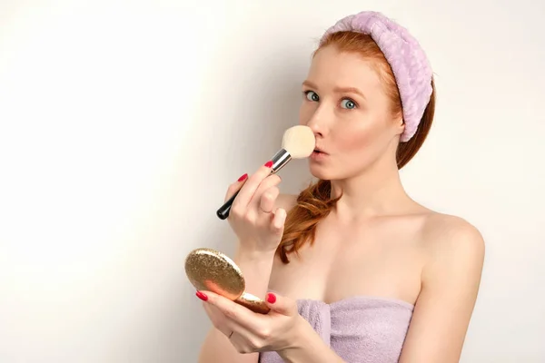 Beautiful redhead girl with clean skin in a towel powders a spout looking at the camera on a white background — Stock fotografie