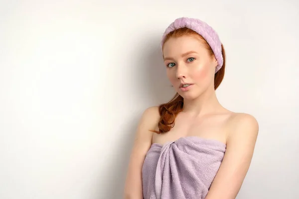 Beautiful red-haired girl with clean skin stands on a white background in a lilac towel. — Stockfoto