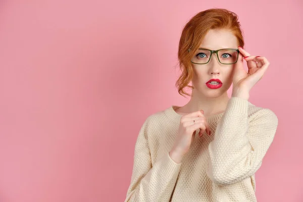 Beautiful redhead girl with red lipstick and manicure stands on a pink background and adjusting his glasses, looking at the camera — Stockfoto