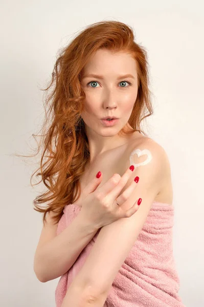 Red-haired girl with clean skin in a towel, draws a heart from a cream on her shoulder with her finger, lips extended — Stock fotografie