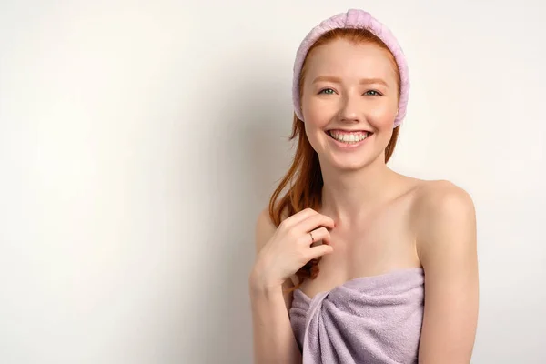 Redhead girl with clean skin in a towel and a bandage on her head openly smiles on a white background — Stock fotografie