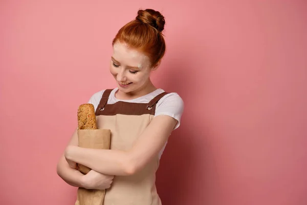 A red-haired girl with gathered hair, in an apron, stands on a pink background, hugging a bag of bread, bowing her head — Stockfoto