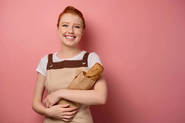 Redhead girl with gathered hair, in an apron, with flour on her face, smiling, holding a paper bag with bread — Stockfoto