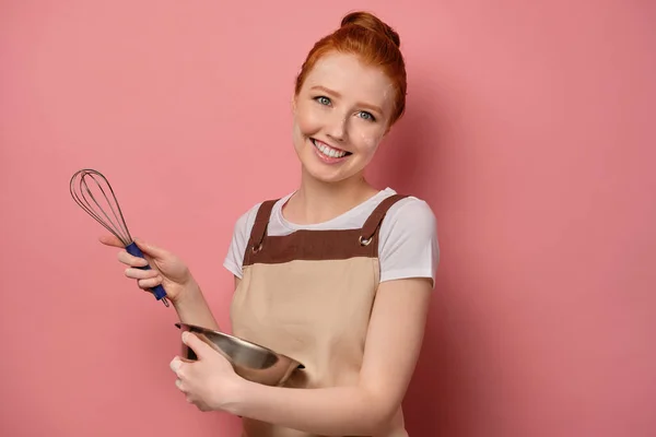 Cute red-haired girl with a high bun in a beige apron and a face in flour, laughs with a bowl and a whisk in hands — Stockfoto
