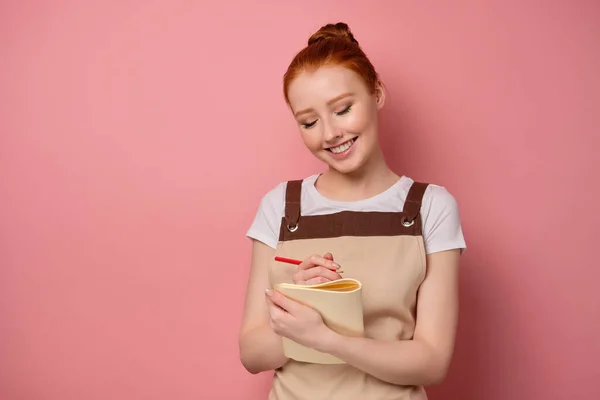 Red-haired girl in an apron with collected hair is standing on a pink background with a notepad smiling and closing her eyes. — Stockfoto